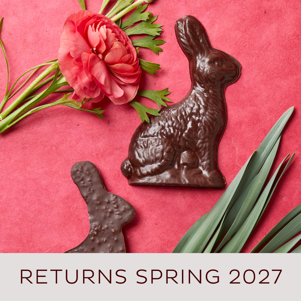 One chocolate rabbit shown in profile is face up and one is face down displaying the bumpy puffed crisps mixed into the chocolate. They are displayed on a red background with a red ranunculus bloom. A badge says: Easter Bunny Sales Benefit The Atlanta Community Food Bank!