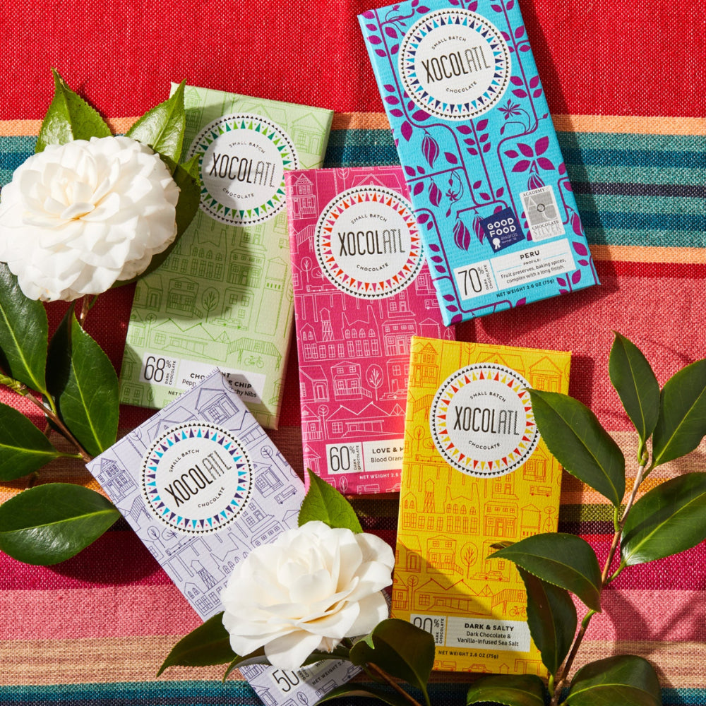5  Xocolatl chocolate bars wrapped in light green, hot pink, yellow, lavender, and teal blue wrappers lying with white flowers on colorful fabric.