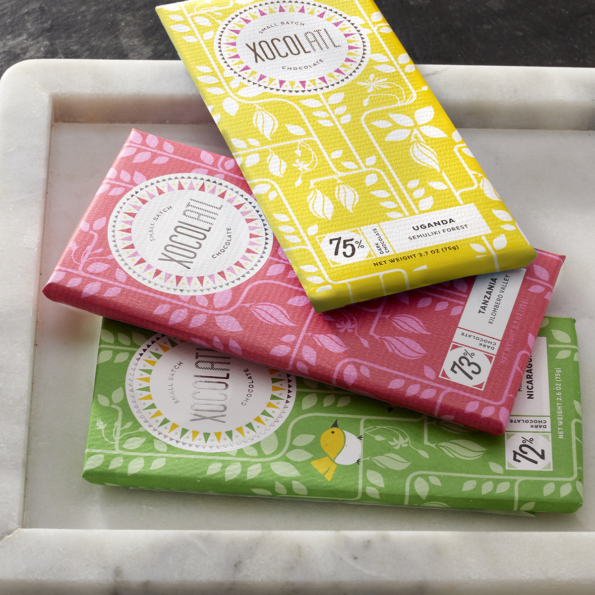 3 colorfully wrapped Xocolatl chocolate bars fanned out on a white marble tray: bright yellow Uganda 75%, cherry red Tanzania 73% and grass green Nicaragua 72%.