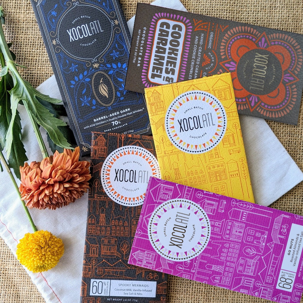 Five Xocolatl chocolate bars in fall wrappers (Cookies & Caramel, Barrel-Aged Dark, Dark & Salty, Spooky Mermaids and Go Nuts!) on burlap with orange and yellow flowers.