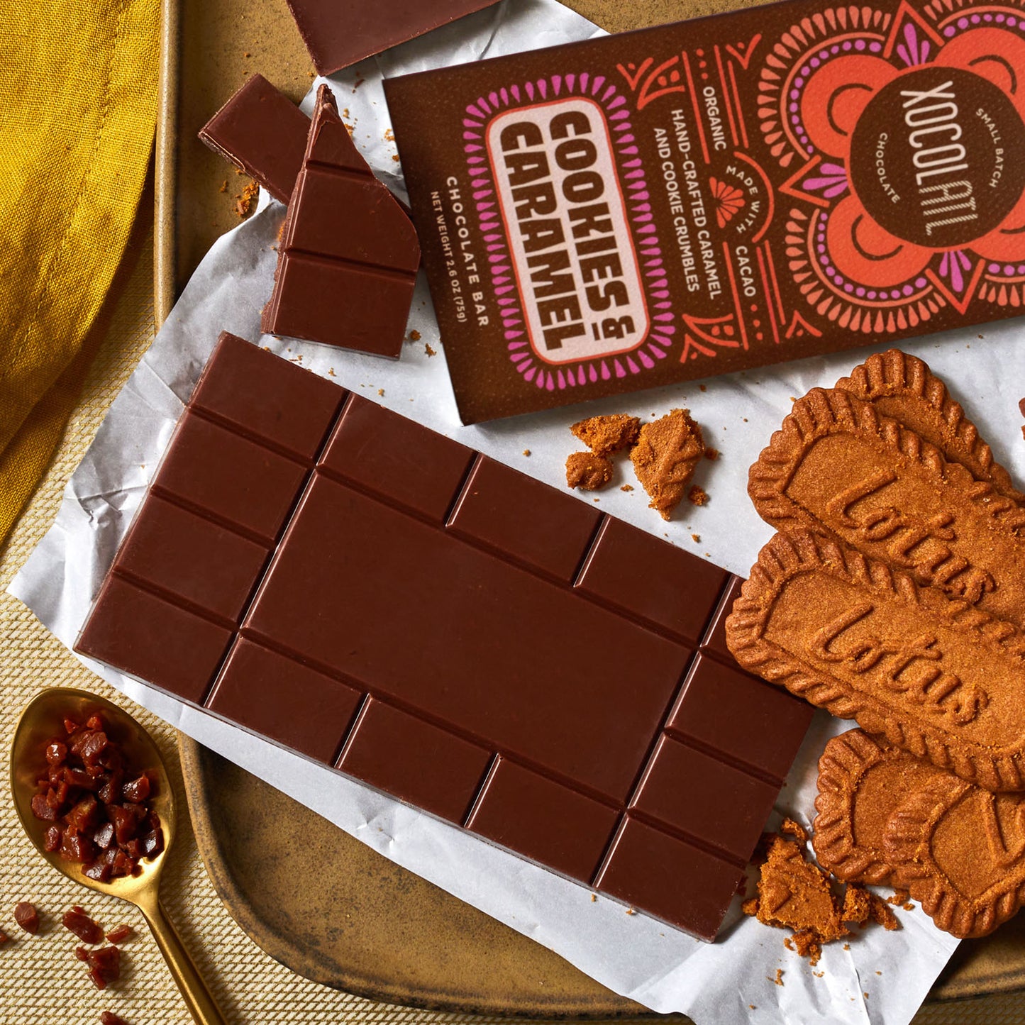 An unwrapped Xocolatl chocolate bar of the flavor "Cookies & Caramel", lying on a tray with opened Biscoff cookies, a spoonful of chopped caramel chunks and the bar wrapper that whimsically reads, "Cookies & Caramel".