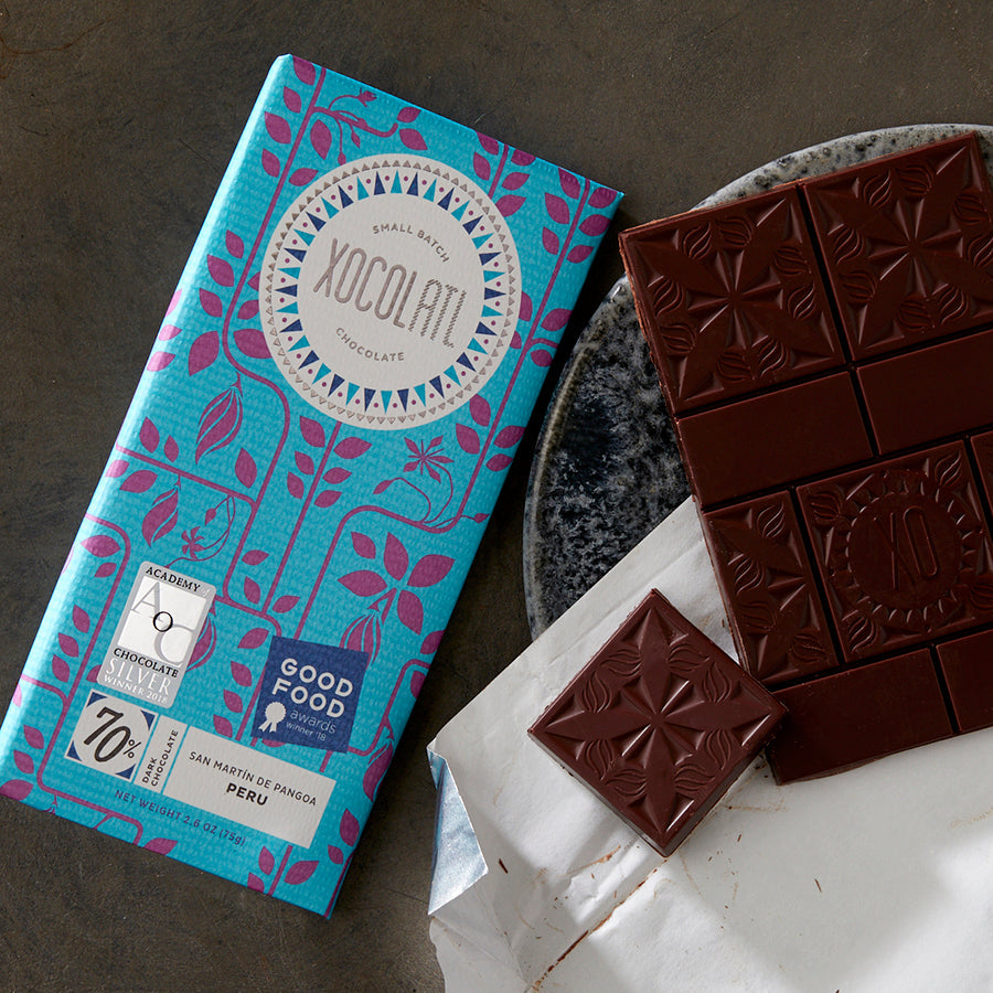 A teal blue Xocolatl chocolate bar of the flavor 70% Single Origin Peru beside an unwrapped and partially eaten chocolate bar on a grey, stone plate.