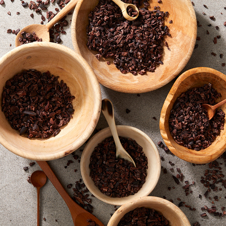 Wooden bowls filled with roasted organic cacao nibs, with wooden spoons and scattered nibs on the surface.