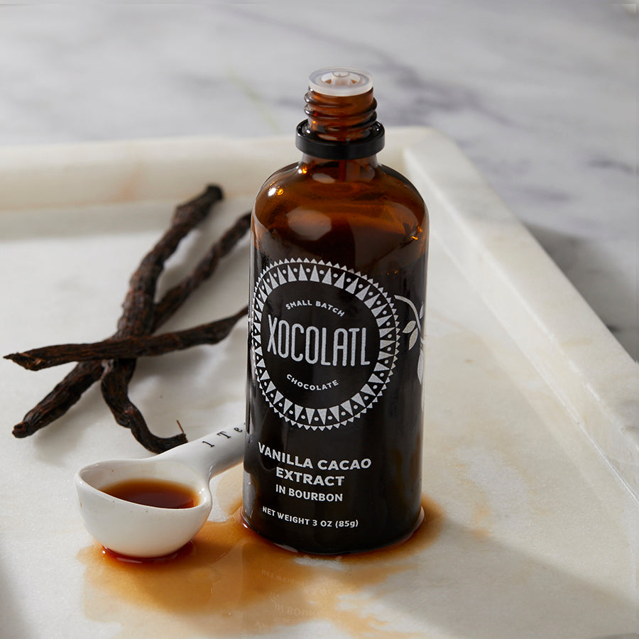 A bottle of XOCOLATL vanilla cacao extract in bourbon, with a vanilla pod and a small measuring spoon on a marble surface.