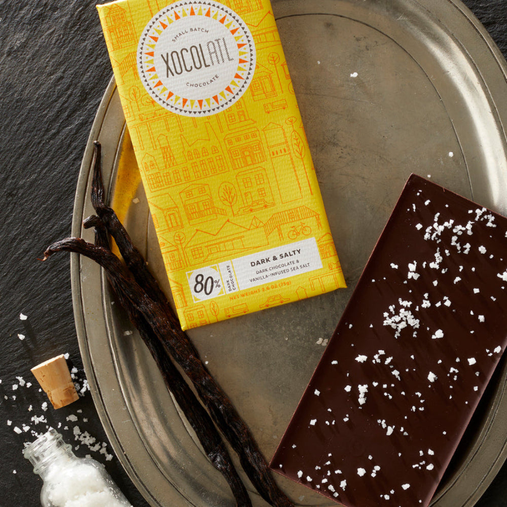 A Xocolatl Chocolate bar in a yellow wrapper, with an unwrapped chocolate bar showing white sea salt flakes sprinkled on it, lie on a silver tray with vanilla beans.