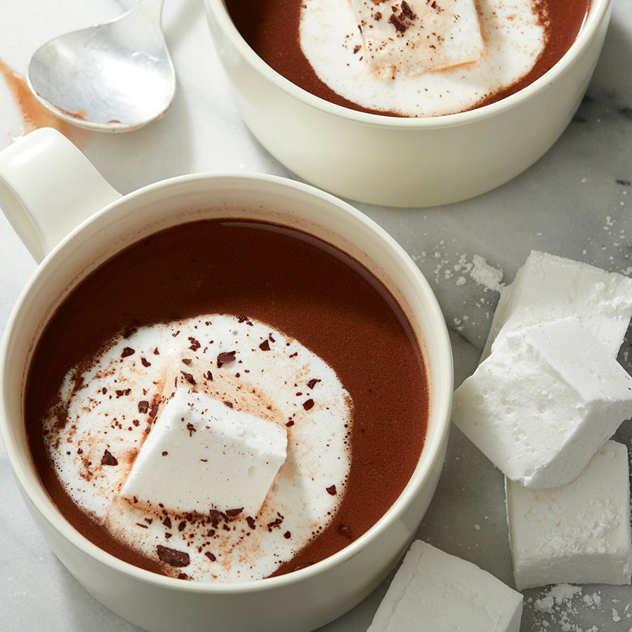 Two white mugs filled with drinking chocolate, topped with whipped cream and cocoa powder, with marshmallows on the side.